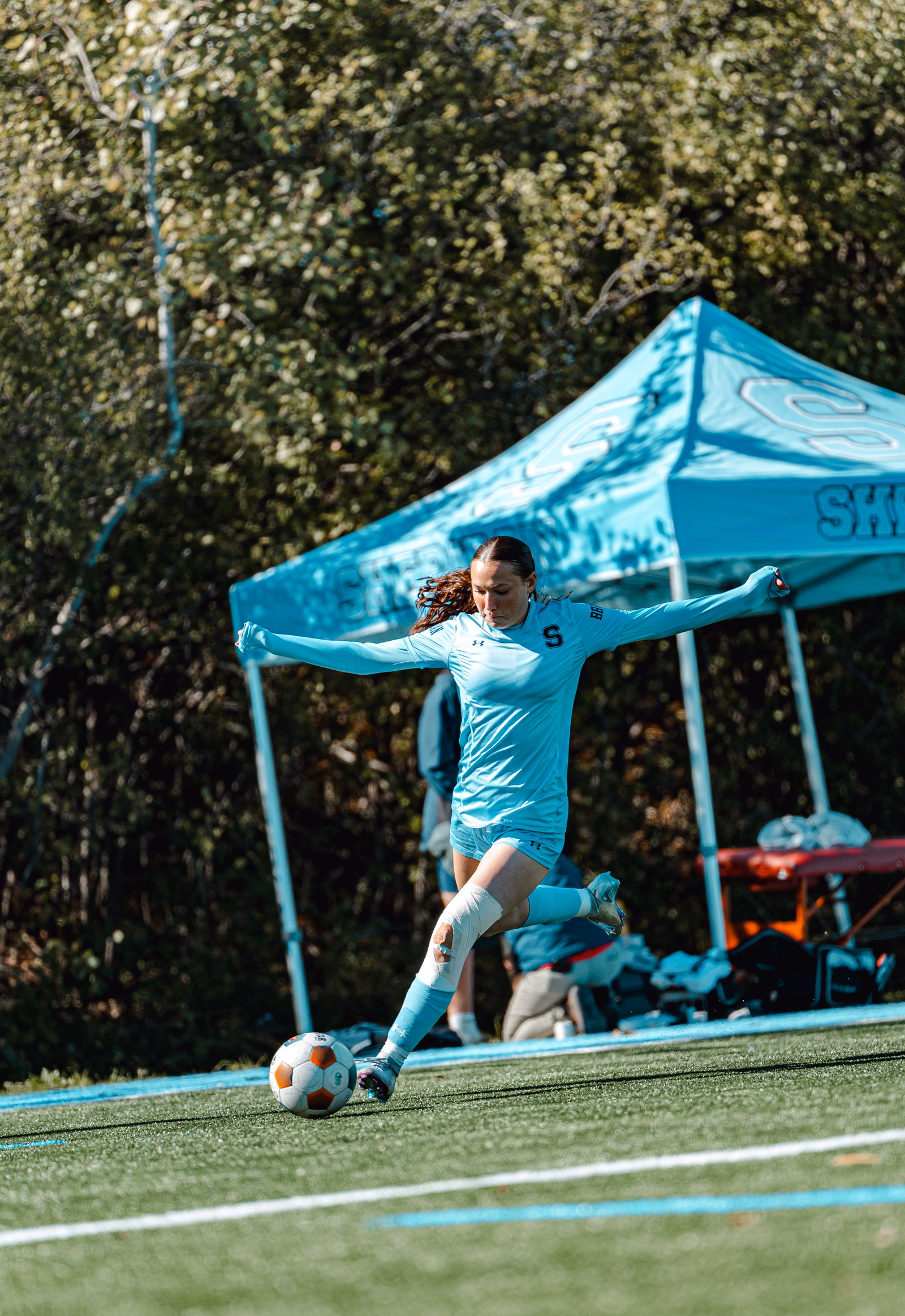 Football player in a turquoise kit striking the ball beside a matching team canopy