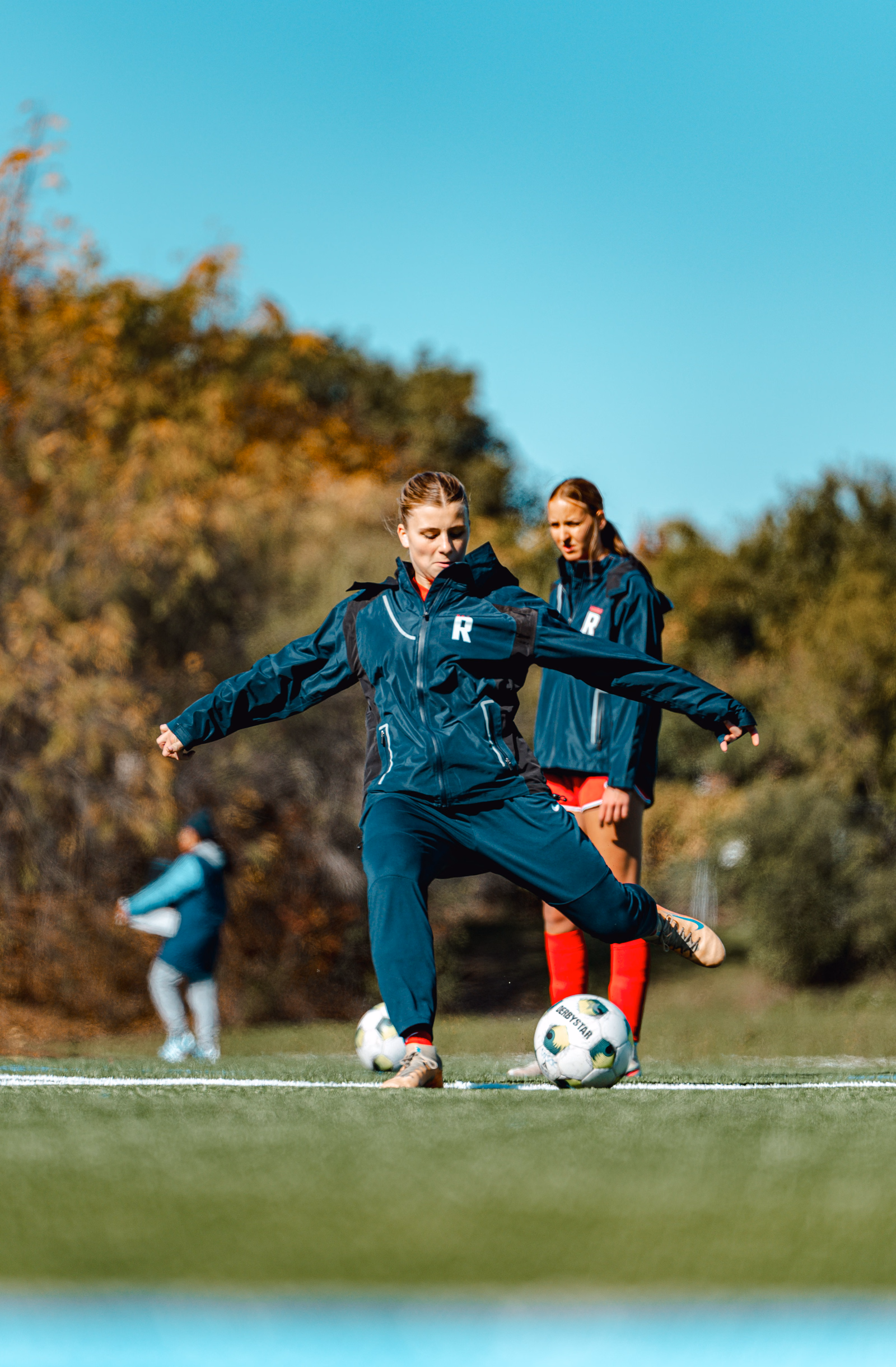 Football player in a teal tracksuit practising a kick during warm-ups